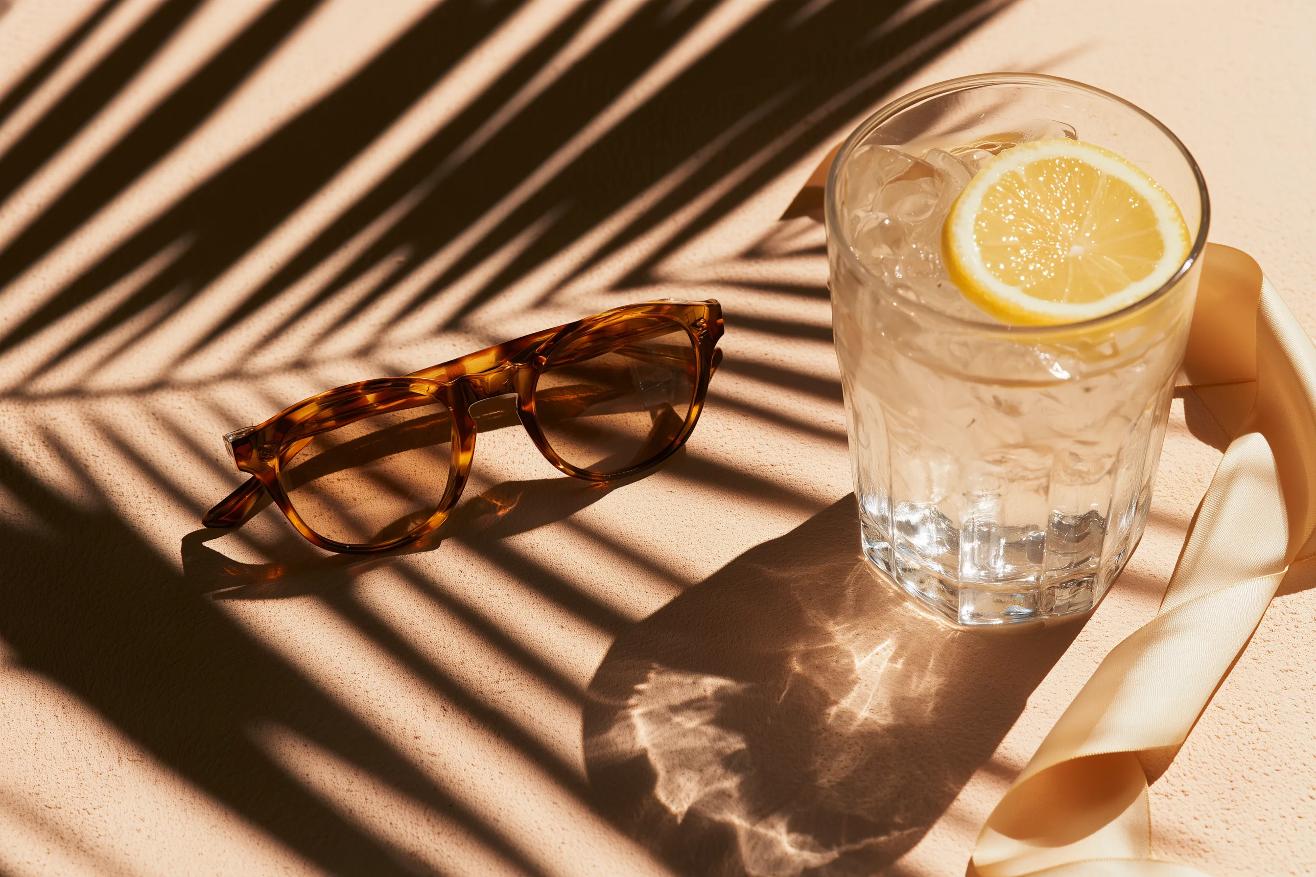 Tortoiseshell sunglasses and a glass of iced water with a lemon slice on a blush surface, striped with palm-leaf shadows in bright sun